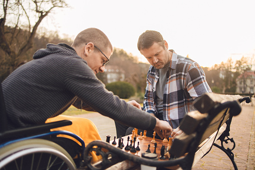Disabled man in the wheelchair spending time in the public park during late autumn season covered with blanket, playing a game of chess and having good times with his friend who is sitting on the bench next to him.