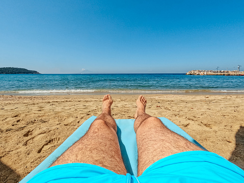 Young caucasian man with hat lying on beach at sea. My son was enjoying the sun while he was relaxing on the sand near the ocean. Water waves are coming from the blue water towards him. Sunbathing and lying lazy in nature is fun to do when you are on vacation in Portugal.