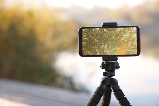 Tripod with smartphone near river. Space for text