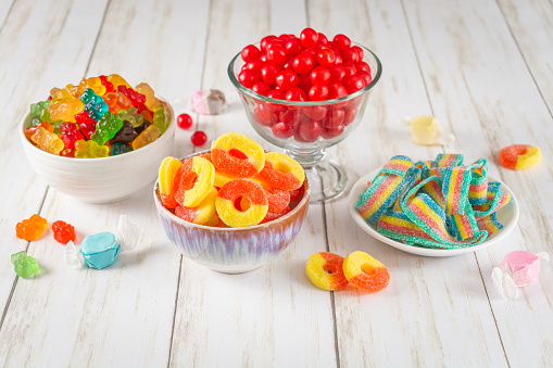 This is a bright and airy photo of a white wooden table with colorful candies on it, including gummy bears, cherry sours, peach rings, and rainbow belts.