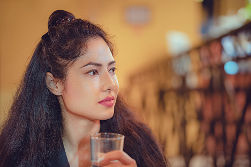 An Asian/Indian young woman drinks water while sitting relaxed in a cafe. She has a half-bun hairstyle in her long hair and wears a black coat.