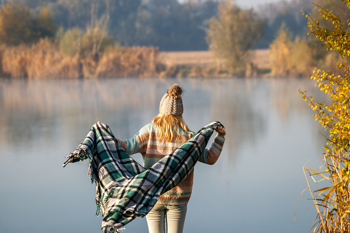 Woman with knit hat is wrapping herself in blanket at lake. Autumn camping in nature
