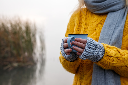 Woman drinking hot drink from mug in cold autumn weather outdoors. Female hands with knitted fingerless glove