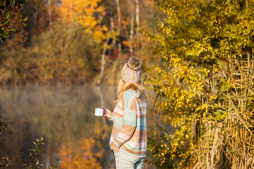 Woman with cardigan sweater and knit hat relaxing with hot drink next to lake at autumn cold morning