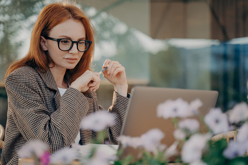 Young focused red-haired business woman works online at outdoor cafe, female in eyeglasses looking at laptop screen with pen in hands during remote work or distant learning at coffee shop