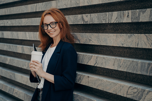 Outdoor shot of glad redhead European woman drinks takeaway coffee holds digital electronic device wears transparent glasses formal clothes stands near marble wall blank space for your text.