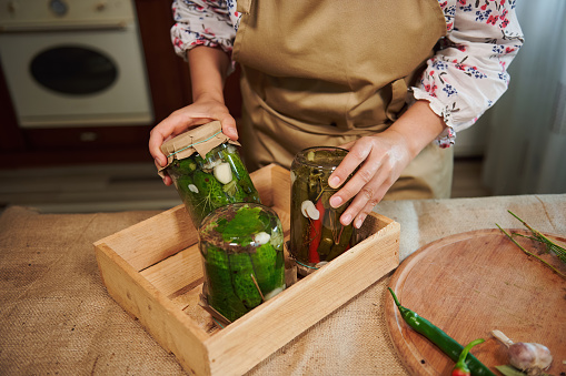 Details: A housewife's hands put canned fermented pickles and pickled chili peppers with vinegar brine upside down on a wooden box in a home kitchen. Preparing homemade preserved for the winter