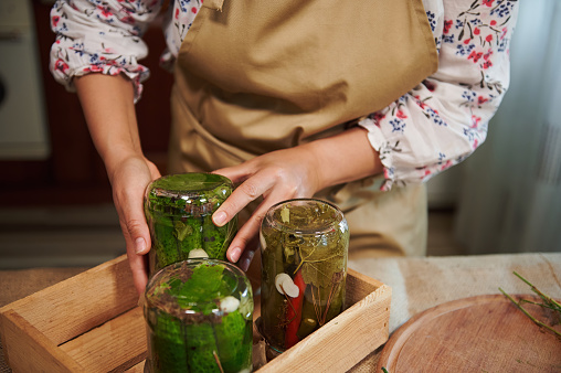 Cropped image of a woman, housewife wearing a beige chef's apron, putting jars of canned vegetables lid down on a wooden crate. Still life with homemade fermented vegetables. Close-up.