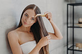 Young brunette woman brushes hair with comb after taking shower and applying hair care mask, wears minimal makeup, has healthy glowing skin after hygienic procedures, stands wrapped in towel