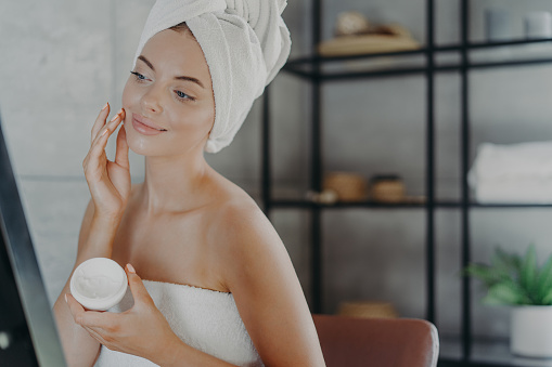 Winsome pleased young European woman puts cream on face, looks at her reflection in mirror, wrapped in bath towel, has anti aging procedure after showering for healthy skin. Skin care concept