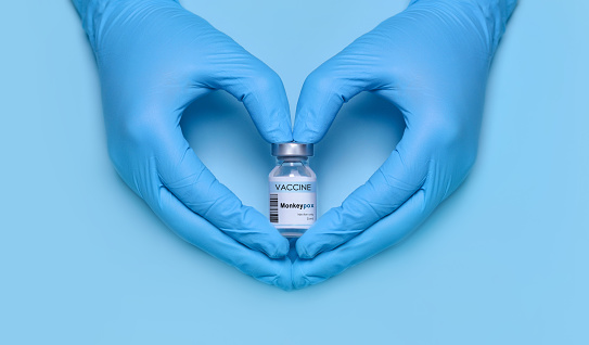 Doctor's hands in protective medical gloves hold a bottle of Monkeypox vaccine in a heart-shaped gesture on a blue background. Vaccination. Immunization. Fighting the pandemic. Monkeypox 2022 virus.