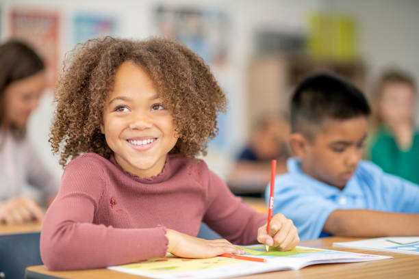 Elementary Student Writing in Class A female Elementary student of African decent, sits at her desk in class as she works on a writing assignment. She is dressed casually and is looking up from her books to smile. Her peers can be seen seated around her in the background at other desks, working away independently as well. elementary student stock pictures, royalty-free photos & images