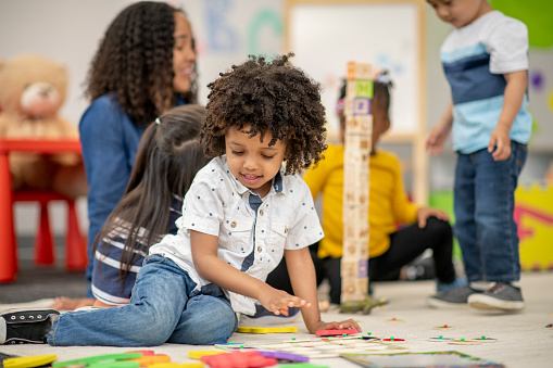 A young Kindergarten student of African decent, plays on the floor among his peers as he focuses on his task.  He is dressed casually and playing with colorful blocks as his peers build a tower with their teacher in the background.