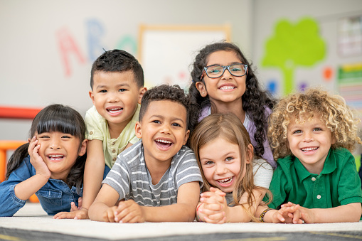 A small group of 6 Kindergarten students lay stacked on top of one another, on their classroom floor, as they pose for a portrait.  They are each dressed casually and are smiling for the photo.