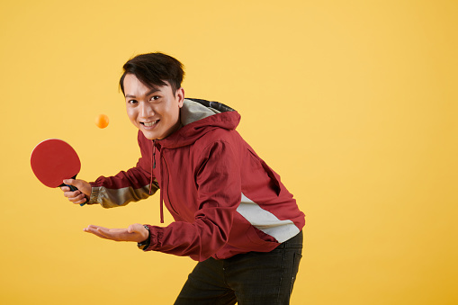 Smiling young Asian man playing table tennis, isolated on yellow