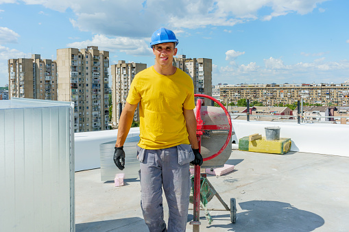 A young Caucasian male construction worker is standing on the roof of a building under construction and pulling a cement mixer behind him.