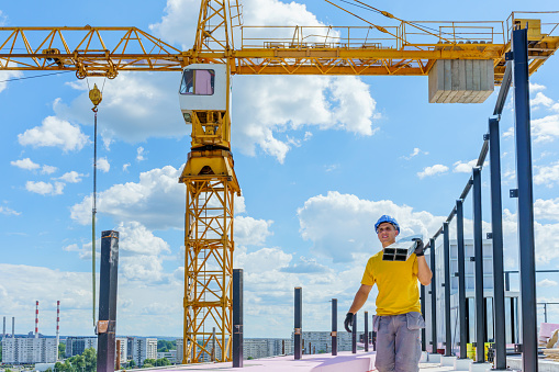 A young Caucasian male construction worker is walking across the roof of a building, carrying metal components, with a construction crane behind him.