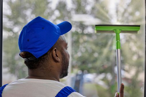 Photo from the back of a strong African-American man in a white T-shirt and a blue cap with a window cleaning brush in his hand. A black guy washes a fogged office window