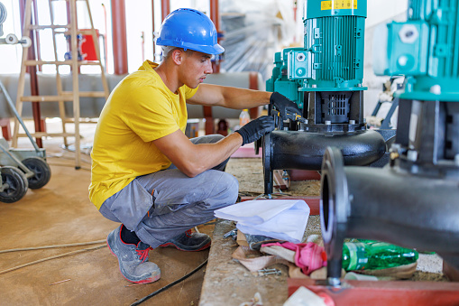 A young Caucasian heating plant worker wearing a hardhat is in a squatting position next to one of the machines, performing a quality check.