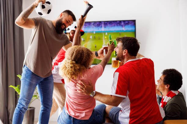 Friends cheering, making a toast and drinking beer while watching football on TV stock photo