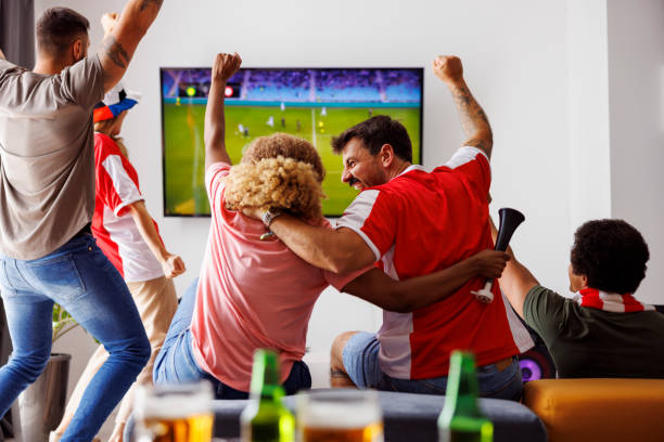 Friends cheering while watching football on TV Group of young friends having fun watching football match on TV, drinking beer and cheering; football fans watching game at home celebrating after their team scoring a goal watching stock pictures, royalty-free photos & images