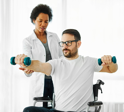 Portrait of a young man in wheelchair exercising lifting weights dumbbell during physical therapy with therapist or doctor at home or in hospital