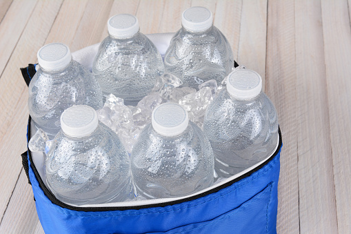 Closeup of six plastic water bottles and ice in a collapsable cooler on a white wood table. Horizontal format with copy space