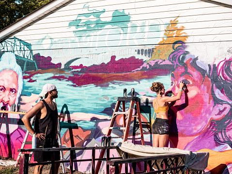 Young painter woman and young painter man artists painting large wall mural she designed. Both dressed in casual work clothes with shorts and summer top. Exterior of large wall of the bungalow house.
