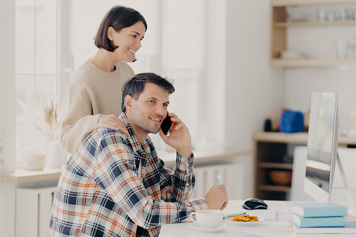 Indoor shot of pleased woman and man busy with discussing domestic expenses, cheerful husband in checkered shirt makes telephone call, caring wife touches shoulders, focuse in monitor of computer