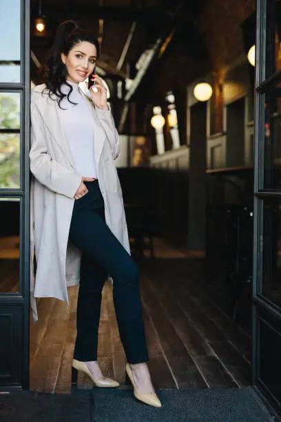 Photo of Stylish young female with dark hair wearing white coat, black trousers and shoes with heels posing into camera while standing against cafe background chatting over phone holding hand in pocket