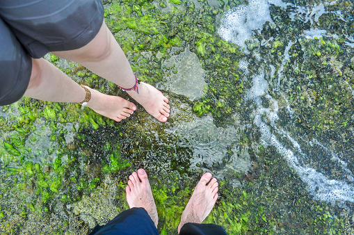 Feet of a young caucasian couple standing on rocks with moss on the beach. Water and leg bracelets