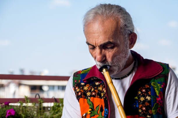 A long, white-haired man in his 60s, with a scruffy beard. He has a unique dress. He is interested in music, has a high life force, positive emotions, an active lifestyle. Wind instrument. He plays the musical instrument by blowing. Opposite laptop video call, train, learning, video conference, master.Model playing musical instrument in front of laptop, portrait shot.