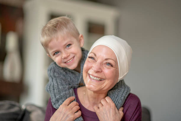 A senior female battling cancer, sits on the sofa as her Grandson reaches around behind her for a hug.  They are both dressed casually and the woman is wearing a headscarf to keep her warm as they snuggle in closely.  Both are smiling and enjoying the quality time together.