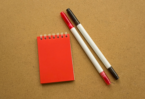 Red notepad for notes with red sheets. Blank space for notes. Mocap. Two felt-tip pens in red and black. On a warm ochre gray background. Holiday calendar notes on notepad. Background for inscriptions