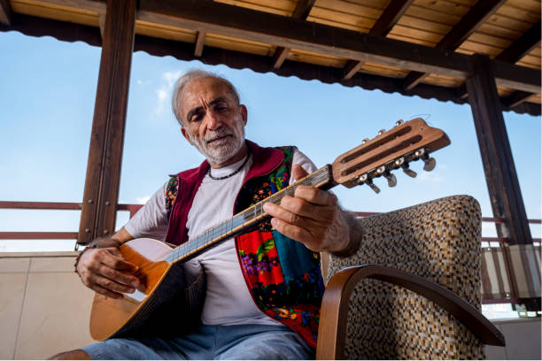 A man in his 60s with long white hair. His scruffy beard is playing baglama. Playing a musical instrument. Active lifestyle with high vitality. Educator, teacher. Model sitting from the waist up and playing a musical instrument. He has a high concentration. He has his own unique clothing.