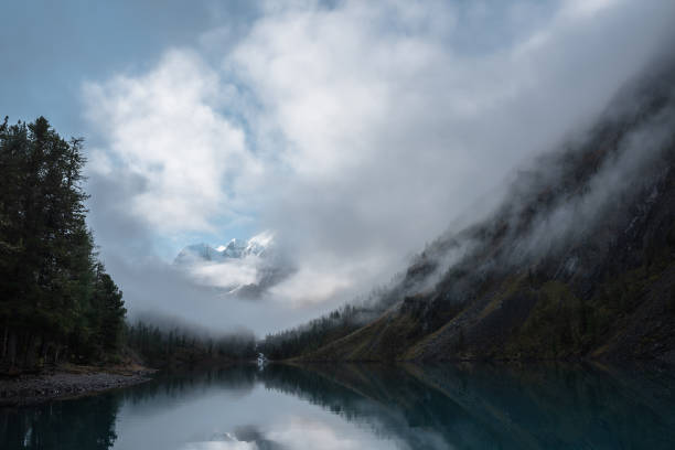 paisaje tranquilo con castillo de nieve en las nubes. el arroyo de montaña fluye desde las colinas del bosque hacia el lago glaciar. montañas nevadas en limpieza de niebla. pequeños árboles de río y coníferas reflejados en un tranquilo lago alpino. - thick snow fotografías e imágenes de stock