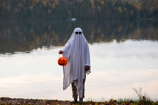 Anonymous person wearing a sheet and sunglasses ghost costume and standing in a sinister fashion in front of a lake at dusk. They are holding an orange pumpkin trick or treat bucket for Halloween.