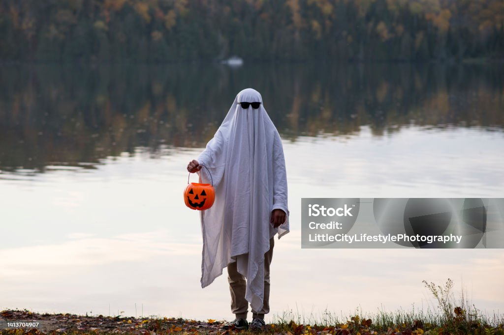 Sinister Ghost Anonymous person wearing a sheet and sunglasses ghost costume and standing in a sinister fashion in front of a lake at dusk. They are holding an orange pumpkin trick or treat bucket for Halloween. Jack O' Lantern Stock Photo