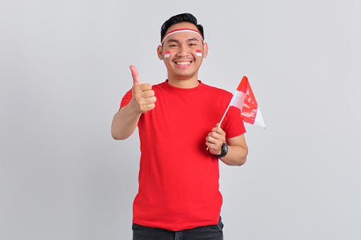 Happy young Asian man holding the Indonesian flag while showing thumbs up isolated on white background. Indonesian independence day concept