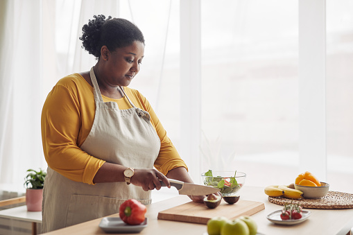 Portrait of young black woman cutting avocado while making salad in sunlit kitchen, copy space
