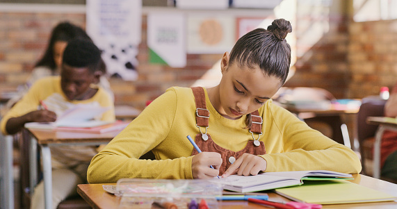 Girl, student or child writing in a book and completing an exam, project or assignment in a classroom at school. Young, serious and concentrating female taking notes in a diary, notebook or journal