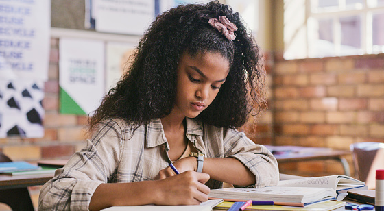 Study, classroom and education for child writing essay or exam at school. Serious teenage student learning and reading from textbook while making notes in her notebook during test or lesson in class