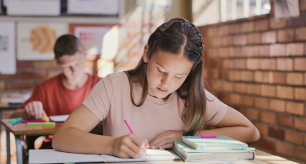 estudio, aula y aprendizaje educativo para niños que escriben ensayos o exámenes en la escuela. adolescente con libros de texto y cuaderno durante una prueba o lección en clase. estudiantes serios sobre la educación - nivel júnior fotografías e imágenes de stock