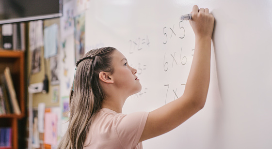 A young clever, smart and intelligent girl writing on a white board in a class a school. A female caucasian student finding a solution for a maths sum in front of a classroom