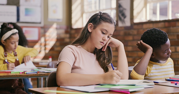 Study, classroom and education for children learning and writing an essay or exam at school. Bored and serious teenage student girl using notebook during a test or educational lesson in diverse class