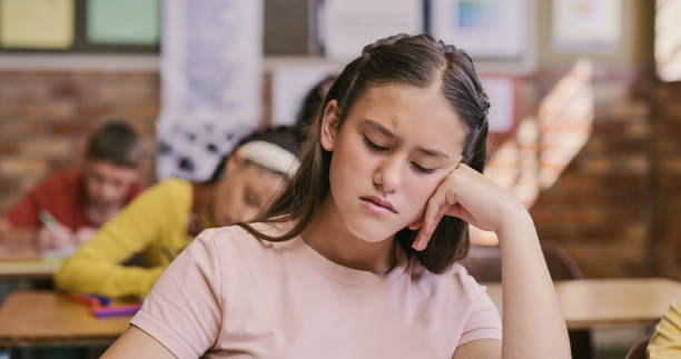 smutna, zdenerwowana i przygnębiona uczennica siedząca w klasie i zmagająca się z edukacją i nauką w swojej szkole. znudzona i zmęczona dziewczyna z adhd lub autyzmem niezdolna do koncentracji podczas lekcji w klasie - learning boredom studying child zdjęcia i obrazy z banku zdjęć