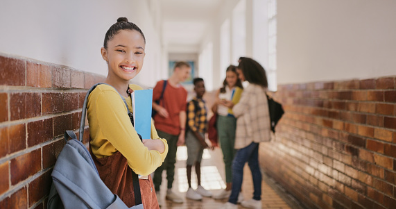Cute young girl excited to be at school and learning with her friends holding her education notebook bag. Portrait of smiling student standing in the hallway with her class mates in the background