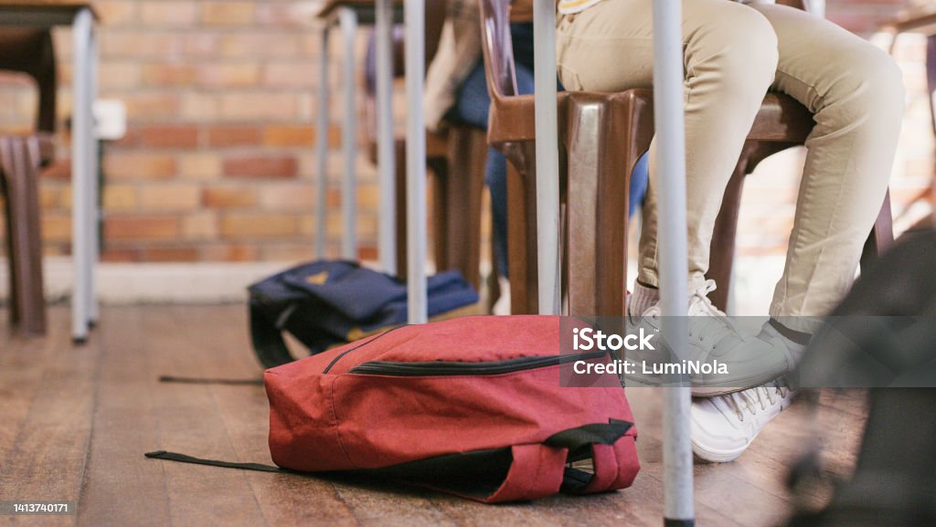 Legs of children sitting in a class at an elementary school learning and getting a quality education. Students learning in a classroom with their backpacks or bags on the floor Classroom Stock Photo