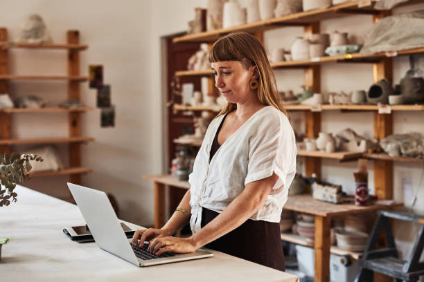 istock Pottery artist tying on a laptop, browsing class or lesson schedule and emailing customers and clients in art studio. Confident artistic potter or creative designer in workshop or hobby craft store 1413740406