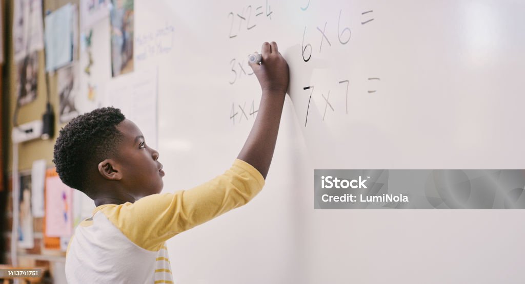 Education, classroom and learning while black student solve a maths equation and write answers on the whiteboard. Smart little school boy doing multiply sum and calculating a solution in class White Background Stock Photo